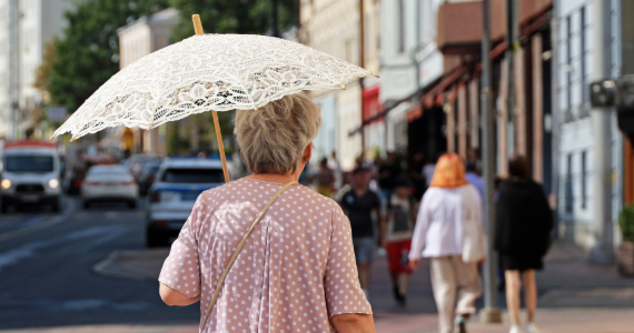 Older woman walks down a city street carrying white parasol to protect from the sun